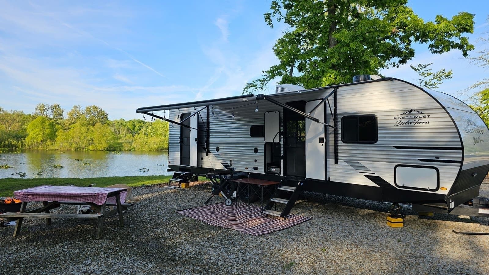 Wanderous travel trailer parked on a gravel pad beside a lake, awning out over an outdoor rug and picnic table