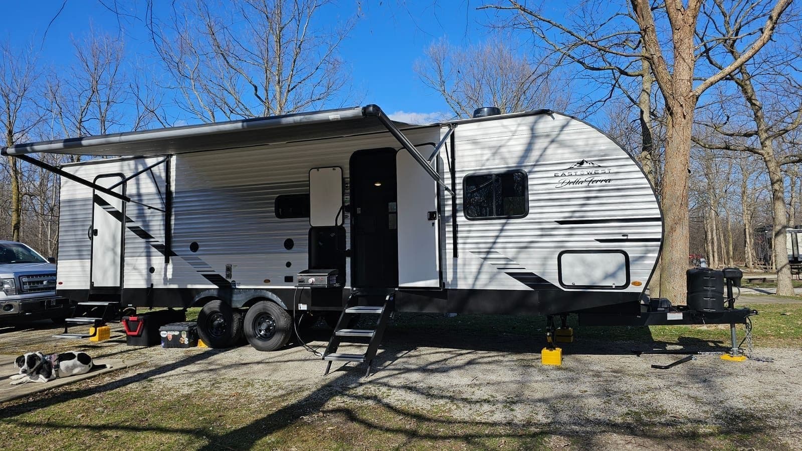 The Great Escape travel trailer on a grass site under bare trees, steps down and yellow leveling blocks in place