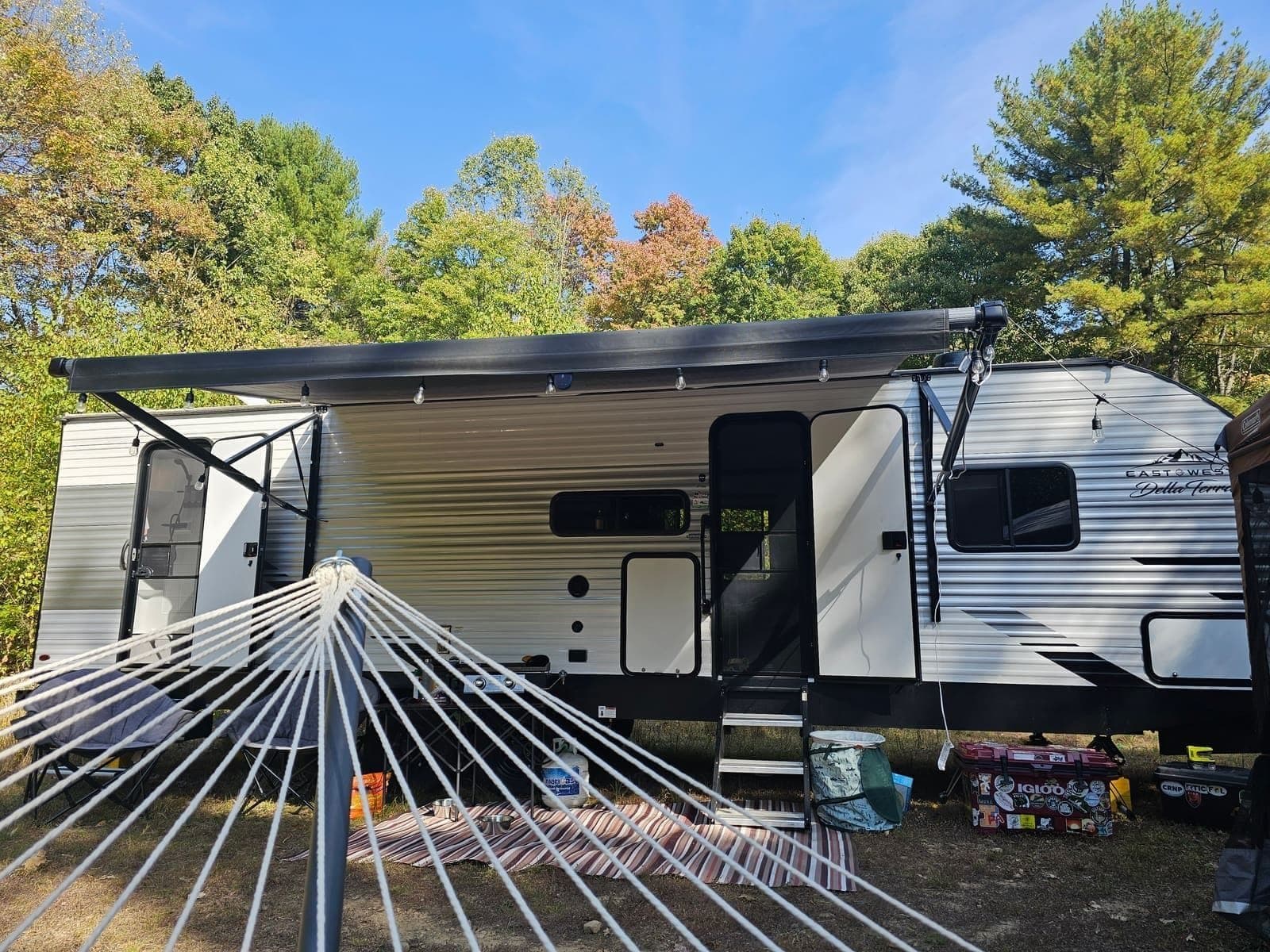 Travel trailer with its awning extended and a striped hammock hung in front, surrounded by autumn woods