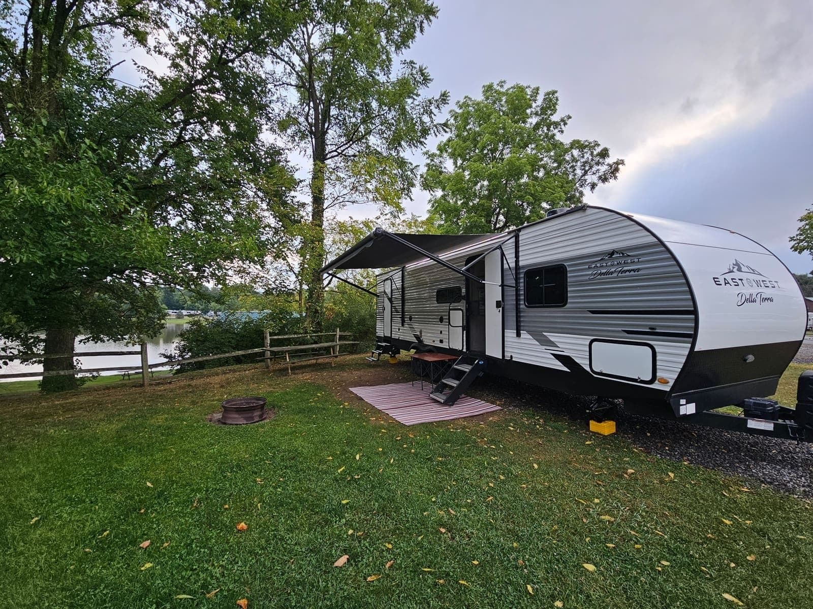 Travel trailer on a shaded grass site with a fire ring in front and water visible through the trees