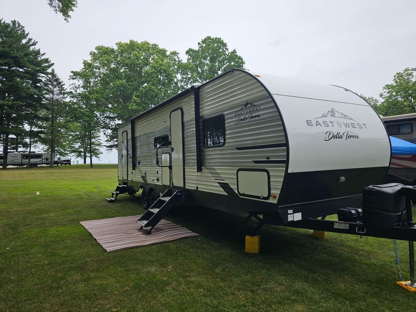 Travel trailer on an open grass site near the treeline with an outdoor rug and steps down