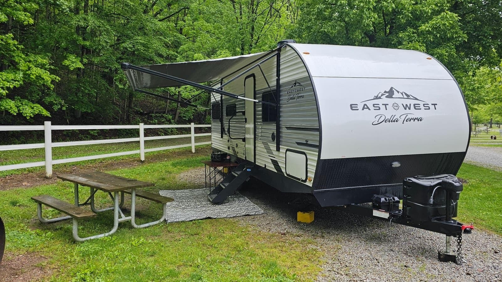 Travel trailer with the awning out on a gravel site beside a white fence, surrounded by fall trees