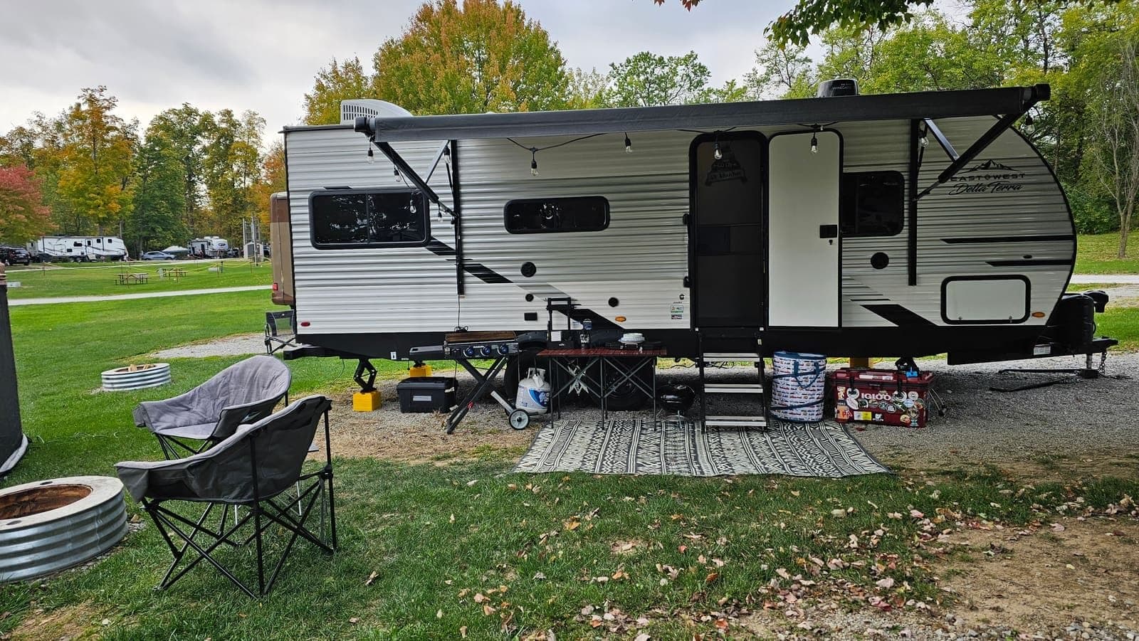 Travel trailer on a wooded grass site with camp chairs and a table of gear set up outside