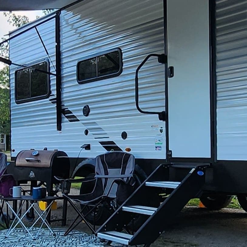 Close view of a travel trailer with a grill and camp chairs set up on the gravel pad beside it